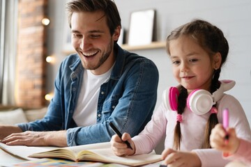 Caring young father helping little daughter with homework. Attentive small schoolgirl studying at home with young daddy