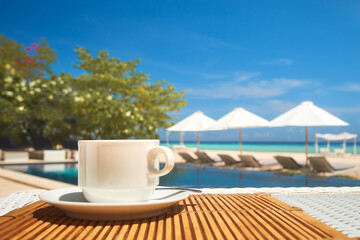 Coffee cup on a table in a restaurant on the beach against the backdrop of a swimming pool and beach umbrellas on a summer day. Focus on the table with a coffee cup. - 724738089