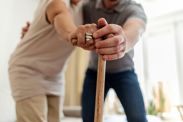 Fototapeta premium Unrecognizable senior woman's hands on walking stick with care worker in background. Male doctor holding a senior patient's hand on a walking stick, special l care concept for Alzheimer's syndrome.