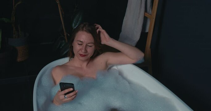 Close-up of attractive young Caucasian blonde woman enjoying bubble bath using smart phone.