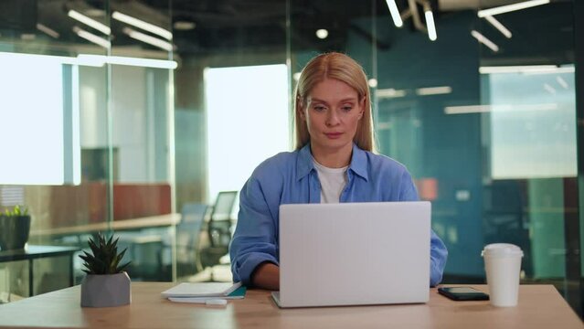 Concentrated woman in blue shirt working on modern laptop by table with personal items and looking aside. Focused female freelancer getting distracted by noise in hall and keeping typing attentively.