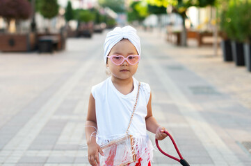 Portrait of a cute little girl with a white turban on her head, pink heart-shaped glasses and a red...