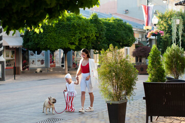 A young stylish mother in white leggings and a white jacket is walking in the city with her little...