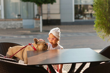 Little cute girl with french bulldog in a chair on the terrace of a cafe in the middle of the city