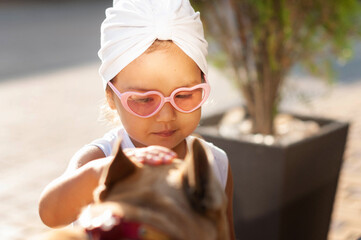 a little girl with a white turban on her head wearing sunglasses is stroking her french bulldog...
