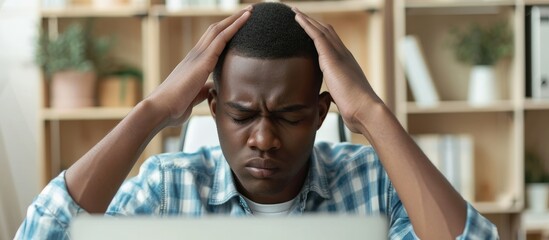 A tired African American office worker, experiencing a migraine and fatigue, seeks rest from working at a laptop in the office, massaging his temples and closing his eyes.
