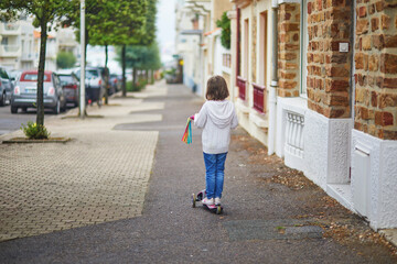 Adorable preschooler girl riding her scooter on a street of Les Sables d'0lonne, Pays de la Loire, France - obrazy, fototapety, plakaty