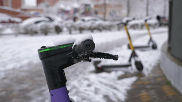 A close-up of an electric scooter covered in snow stands on a snowy road in the city. Rental electric scooters are covered with snow after a snowstorm. Electric scooter handles in the snow.