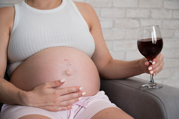 Close-up of the belly of a pregnant woman holding a glass of red wine while sitting on the sofa. Skin rash. 