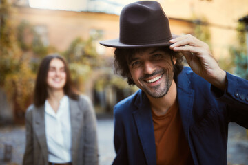 Handsome man donning a stylish hat with charming smile, accompanied by his girlfriend in the background - 724851812