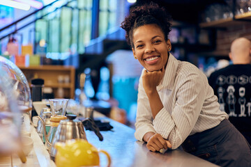 A smiling African female barista, standing behind the bar, working a morning shift.