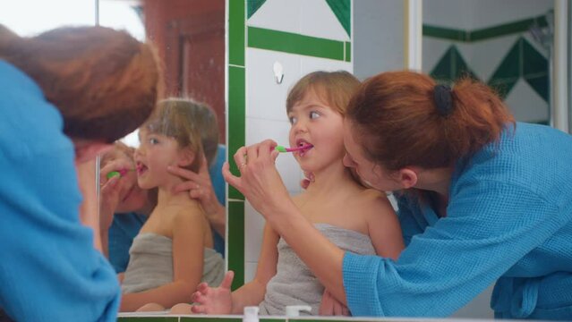 Happy mother is teaching her child to brush teeth patiently at home near mirror