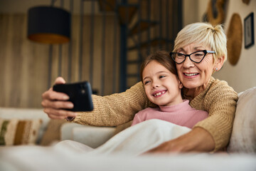 Grandma and her grandchild taking photos with a mobile phone, sitting on the sofa, covered in a blanket. - obrazy, fototapety, plakaty