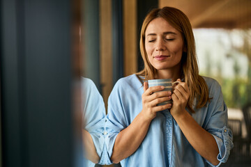 A lovely woman keeping her eyes closed, holding a cup of coffee, standing near the window.