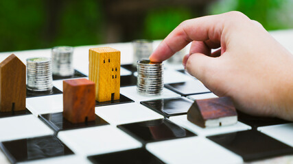 Hand touch stacks of coin money on chess broad with blur wood house model background, Real Estate...