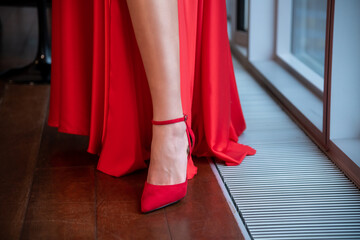 Close-up view of woman wearing red long dress with slip and red high heels standing next to apartment's window. Soft focus. Copy space. Beauty and fashion theme.