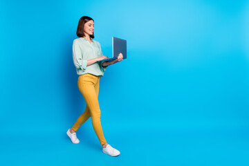 Full size photo of pleasant girl wear turquoise blouse yellow pants walk to empty space look at laptop isolated on blue color background - obrazy, fototapety, plakaty
