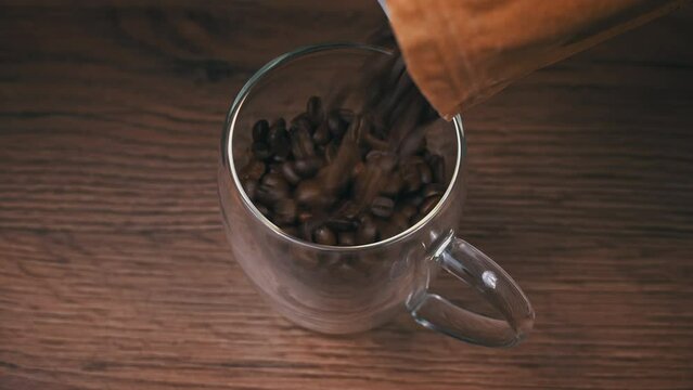 Coffee beans in craft packaging fall into a cup. Roasted aromatic brown coffee beans. Dark roast Arabica coffee beans falling. Roasted aromatic coffee.