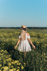 a girl in a hat and a white dress is standing with her back turned in a blooming yellow rapeseed...