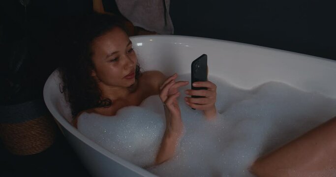 Close-up relaxed happy attractive young African American woman dancing and relaxing in bubble bath, using smartphone.