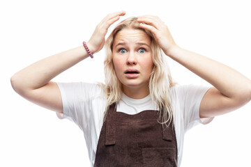 Fototapeta premium The teenage girl raised her hands up in shock. Cute blonde girl in a white T-shirt and brown overalls. Horror and surprise. White background.