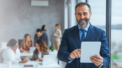Smiling mid aged business man wearing suit standing inside office holding digital tablet. Mature businessman professional holding fintech device looking away thinking or new business ideas solutions