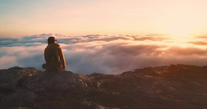 Young hiker girl sits with backpack at mountain top and enjoys beautiful golden sunset