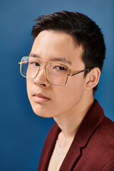 good looking asian teenage boy with glasses in stylish attire looking at camera on blue backdrop