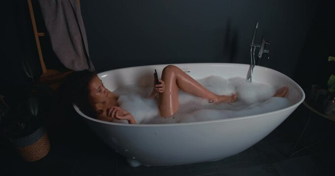 Happy beautiful young woman enjoying relaxing foamy bath and checking social media on smart phone.