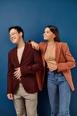 joyful diverse teens in fashionable attires posing on blue backdrop and looking away, friendship day