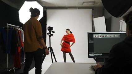 Backstage of model and professional team in the studio. Brunette woman poses for photographer, editor sitting at the desk looking at monitor.