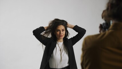 Backstage of model and professional team in the studio. Close up of brunette model in suit posing, photographer taking pics, playful expression.