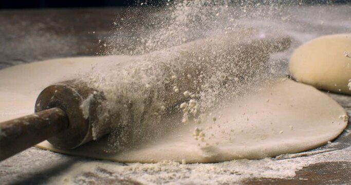 Super slow motion macro of artisan baker chef sprinkles flour dust on rolling pin with raw dough during is making handmade bread, pasta or pizza on rustic wooden table in traditional bakery kitchen.