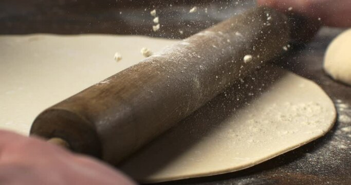 Super slow motion macro of artisan baker is rolling pizza dough with flour dust falling during making homemade traditional recipe italian pizza in bakery and pizzeria restaurant kitchen.