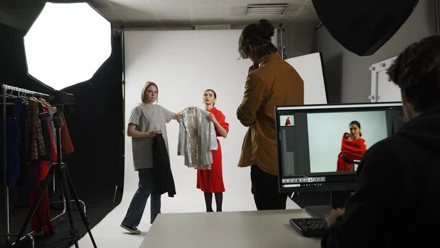Model and production team in the studio. Female posing for photographer, girl assistant chooses clothing, editor at the desktop checking photos.