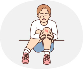 Crying teenager boy sits on ground and holds on to injury on knee after fall during walk. Unfortunate child was injured during sports activities or active games with friends