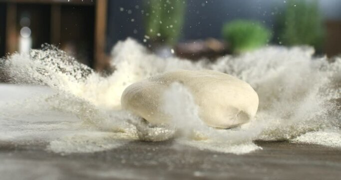 Super slow motion macro of raw loaf of dough falling with flour dust splashes while artisan baker chef making homemade bread, pasta or pizza on rustic wooden table in traditional bakery kitchen.