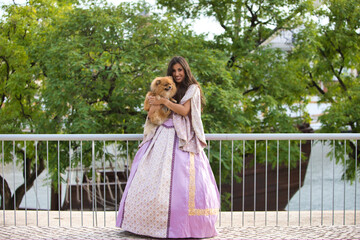 Fototapeta premium Beautiful Latin woman with long curly hair wearing a 15th century dress is holding a pomenarian dog in her arms. The woman is happy with her pet. Dog and pet day concept.