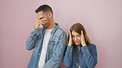 Peeking through fingers, a beautiful couple in denim shirts show a shocked look over isolated pink background, sharing a moment of embarrassing surprise together