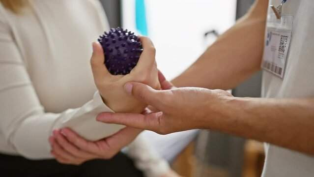 A woman undergoes hand therapy with a spiky ball, assisted by a male therapist in a clinic.