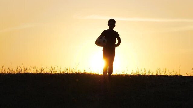 Silhouette of a boy walking down a slope during sunset. The child is holding a soccer ball. Kids dreams, children in sports