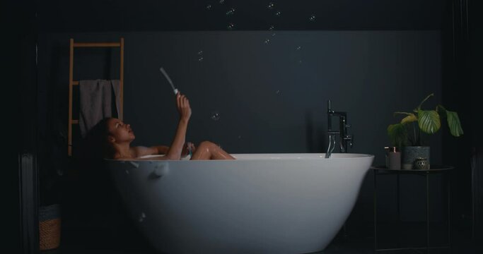 Happy serene relaxed beautiful young woman with curls lying in modern stylish bath having fun blowing soap bubbles.