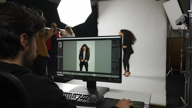 Model and production team in the studio. Full length woman model poses for photographer on set, editor checks photos on monitor.