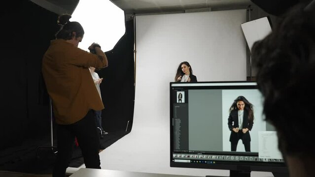 Model and production team in the studio. Full length woman poses for photographer, stylist fixes the look, editor checks photos on monitor.