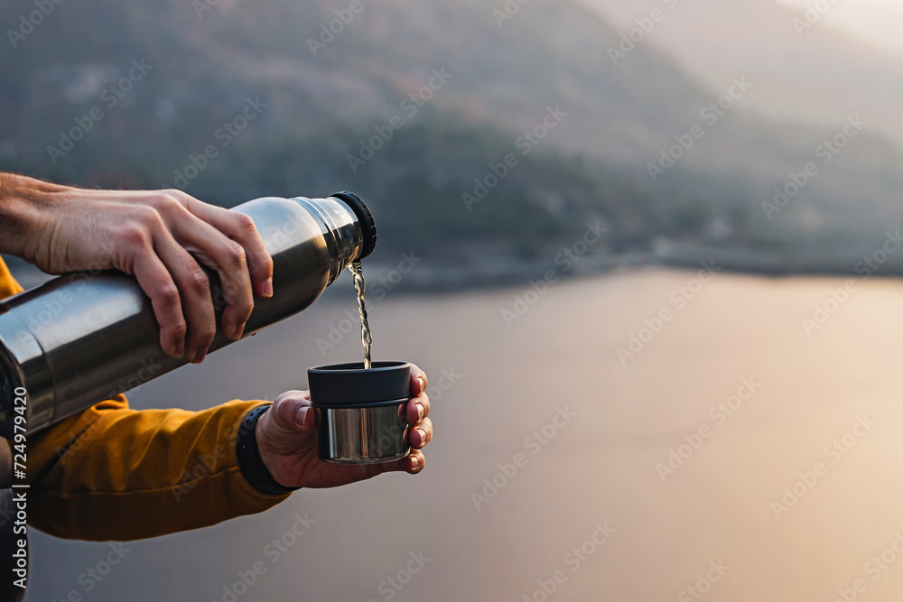 Wall mural man pouring hot tea from thermos outdoors with a beautiful scenic view on the background