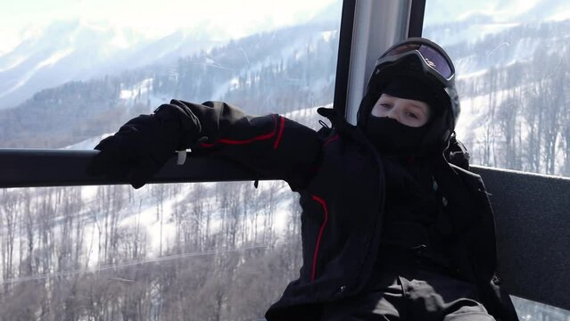 Boy in black shirt, helmet and mask sit in cabin of chairlift, slow motion.