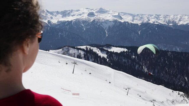 Back of woman in red dress watching on paraplane flying above snow slopes