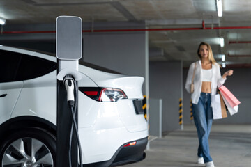 Young woman travel with EV electric car to shopping center parking lot charging in downtown city...