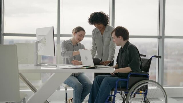 Paralyzed man works with colleagues. Guy in wheelchair has spine trauma discussing working project in office on meeting with multiethnic team group colleagues. Teamwork brainstorming, special needs.