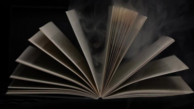 Old mystical book with open pages with magic smoke on black background. Enchanted book with magic light in the dark. Open book lies on the table, illuminated by beam of light among the falling smoke.